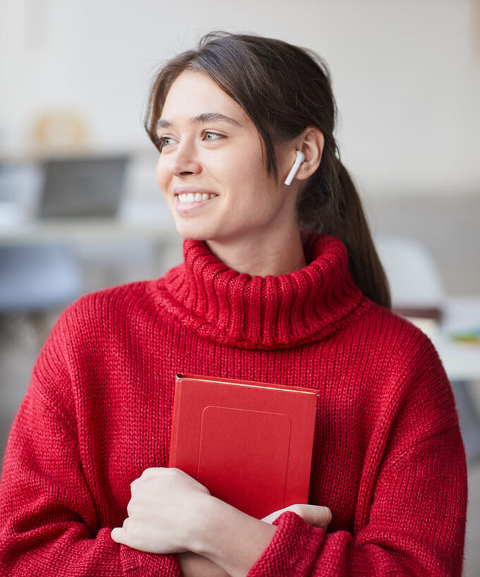 Sprachschule-Lenz-3 Eine junge Frau im roten Pullover hält ein Buch in den Armen und blickt zur Seite.